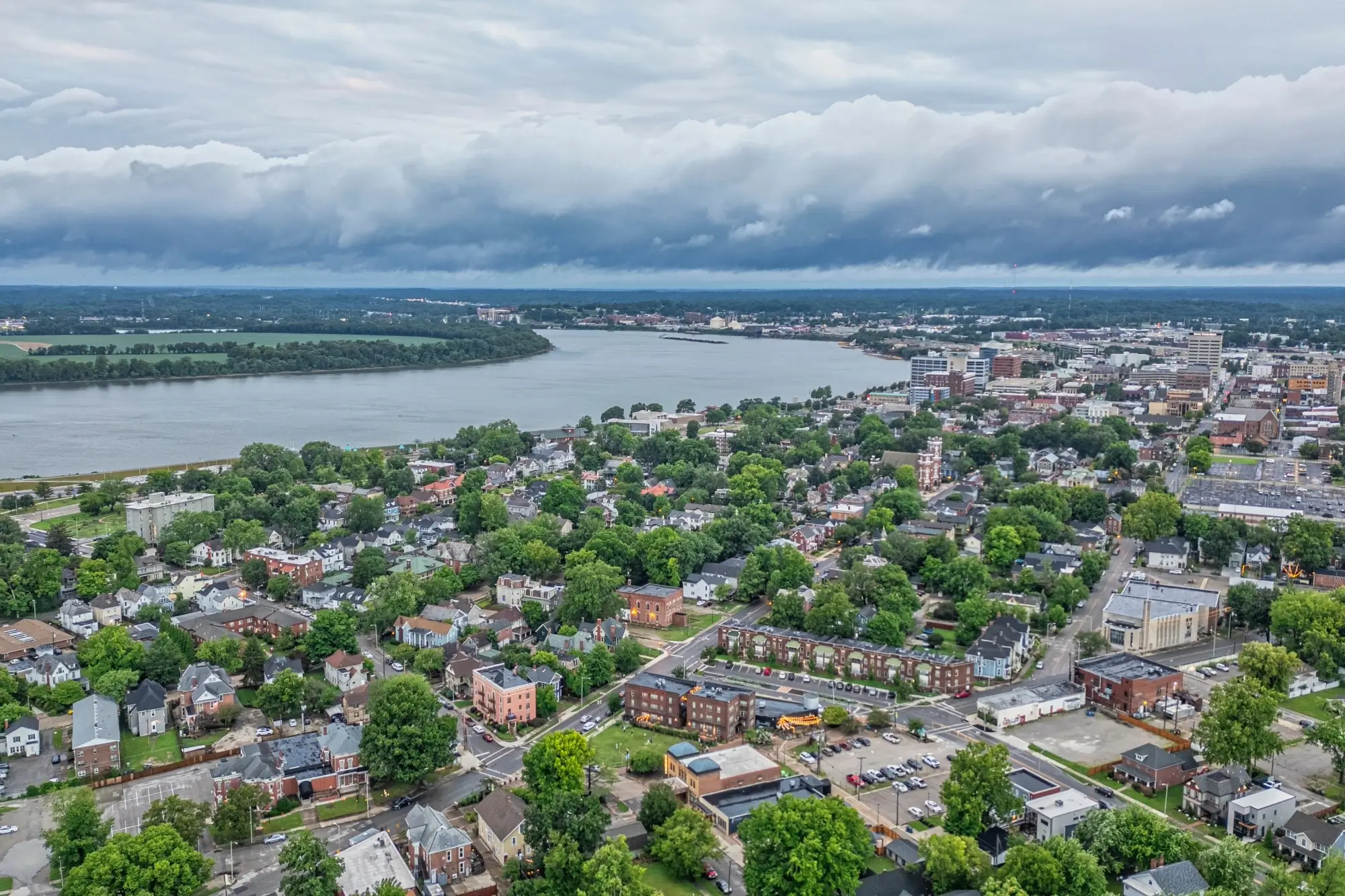  An aerial view of Evansville, Indiana, featuring a mix of historic residential neighborhoods and downtown buildings situated along a wide bend of the Ohio River under a cloudy sky.
